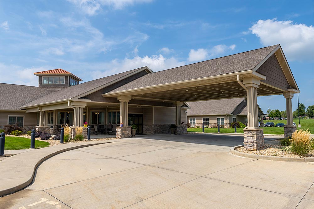 A single-story building with a covered driveway entrance under a blue sky. The senior living facility in Illinois has beige siding, stone accents, and well-maintained landscaping with grass and shrubs.