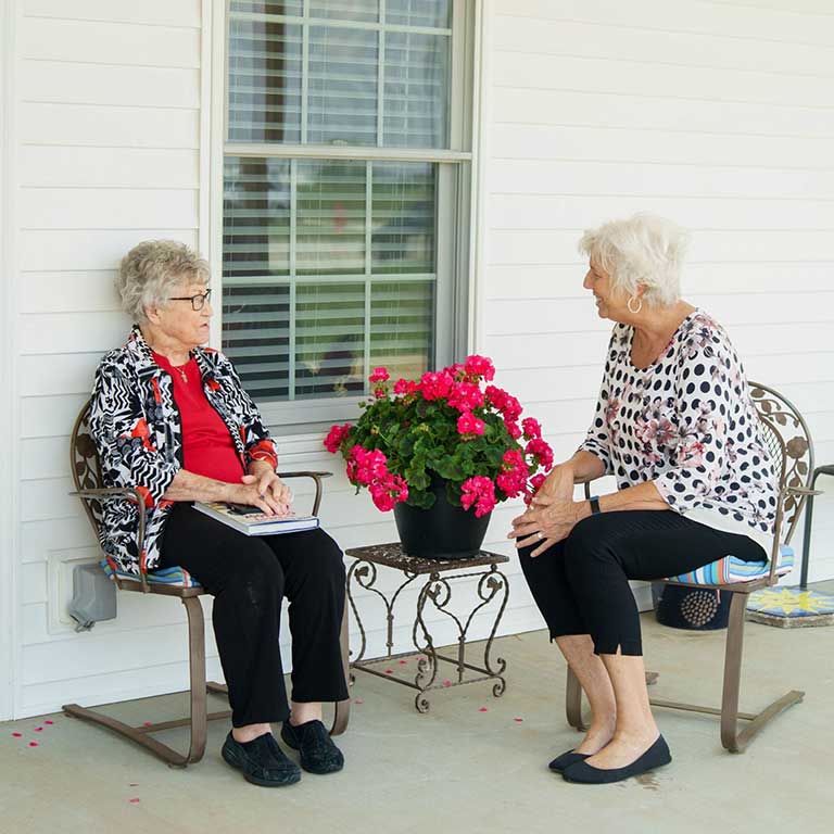 Two elderly women sit and talk on a porch at a senior living facility in Arcola, IL, with a pot of pink flowers on a small table between them.