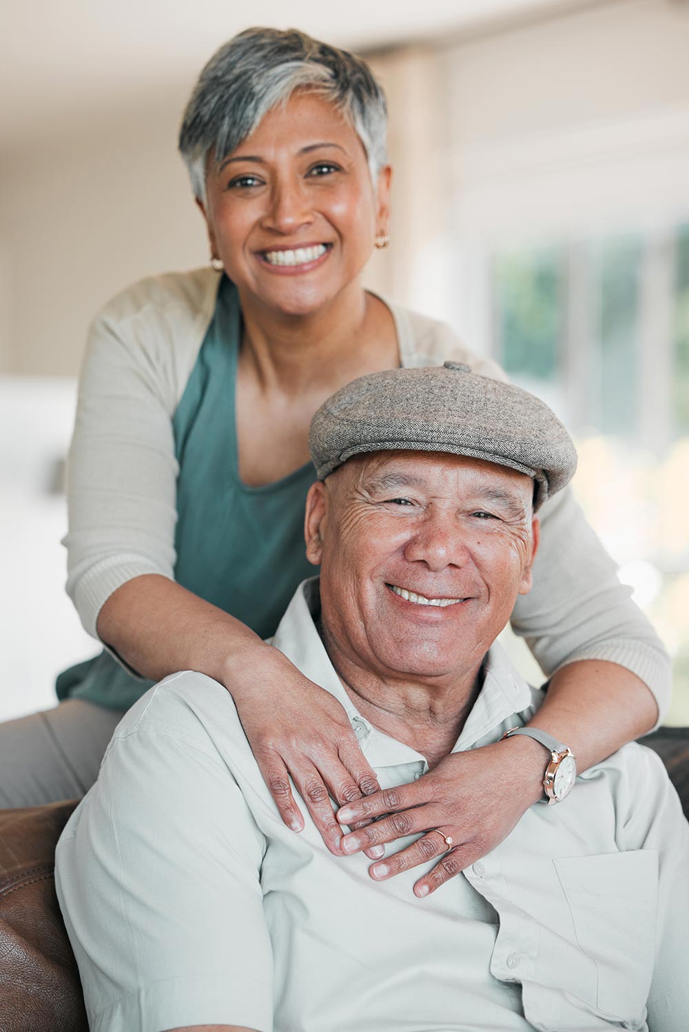 A smiling woman with short gray hair, embodying the warmth of senior living in Illinois, stands behind a man seated on a couch, wearing a cap. Both look at the camera, with the woman's hands gently resting on the man's shoulders.