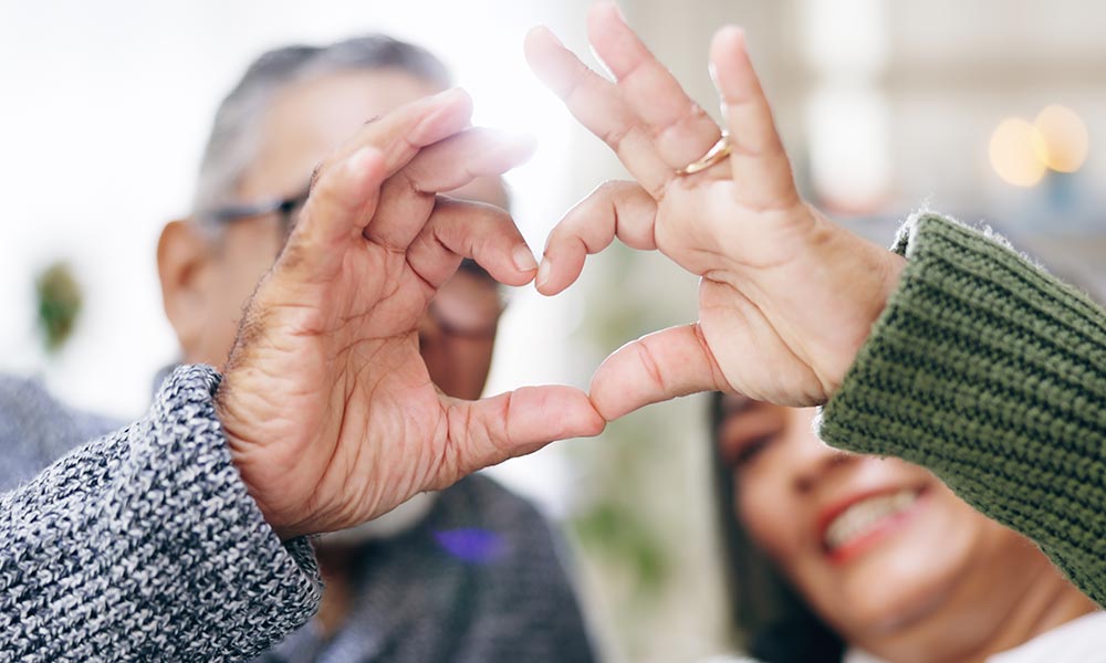 Two elderly individuals in senior living in Illinois are smiling and forming a heart shape with their hands in front of their faces.