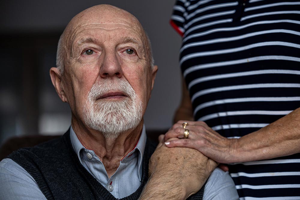 An elderly man with a solemn expression looks ahead while a person wearing a striped shirt gently rests their hands on his shoulder, in a serene senior living community in Illinois.