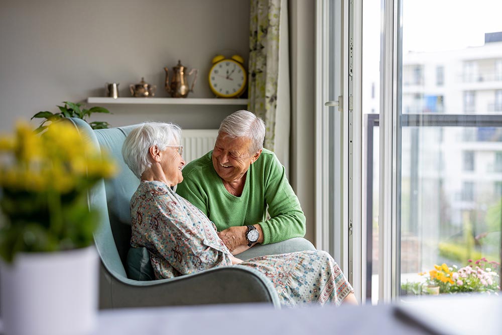 An elderly couple sits and smiles at each other in a cozy living room near a window, enjoying their golden years while embracing the tranquility of senior living in Illinois.