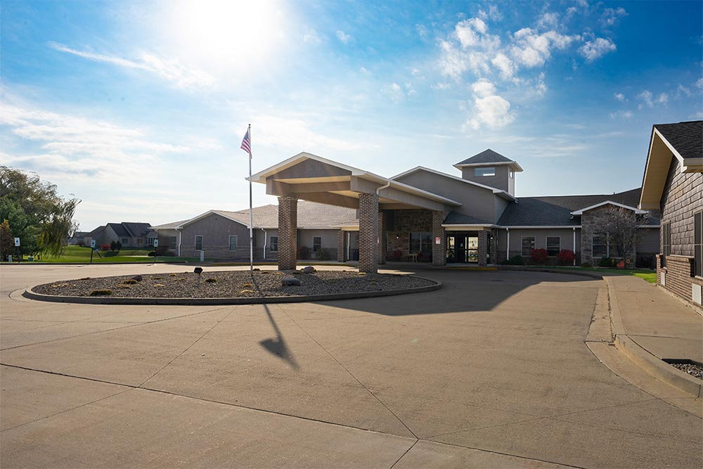 A single-story senior living building in Illinois with a prominent entrance and circular driveway under a clear sky with scattered clouds. An American flag is displayed on a flagpole near the entrance.