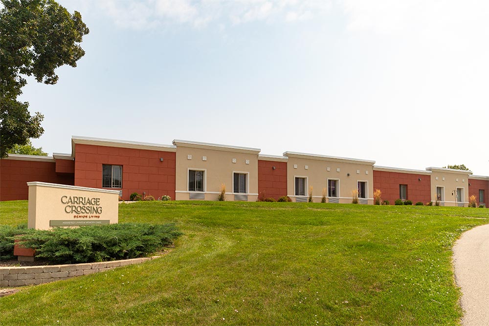 A single-story building with a light yellow and brown facade features a sign that reads "Carriage Crossing Senior Living in Illinois" on a grassy lawn.