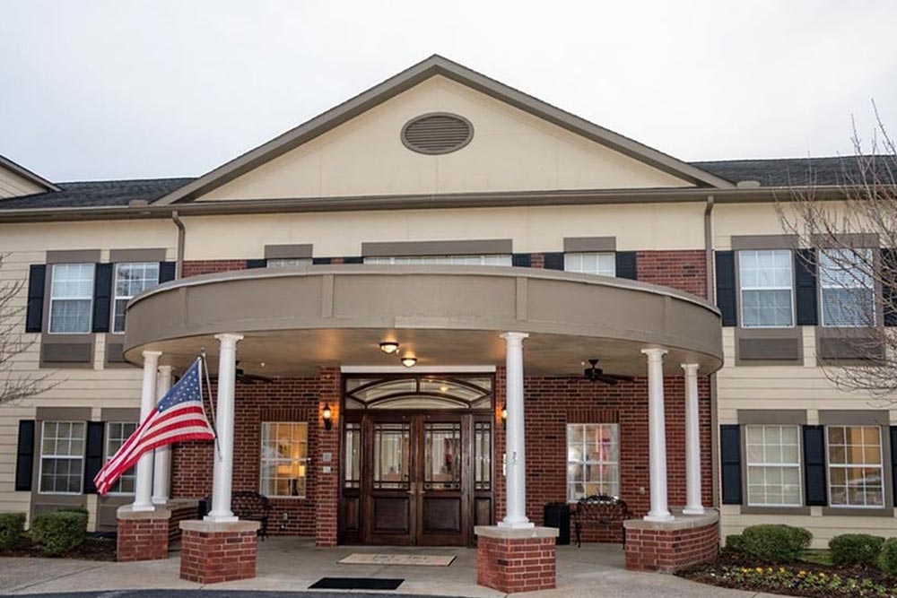 Front view of a two-story senior living building in Illinois with a circular driveway, covered entrance with columns, and an American flag on the left. The facade features a mix of brick and beige siding.