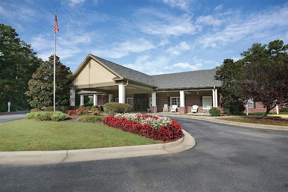 A single-story senior living building in Illinois with a covered entrance, surrounded by landscaped greenery and colorful flowers, an American flag on a pole in front, and a circular driveway.