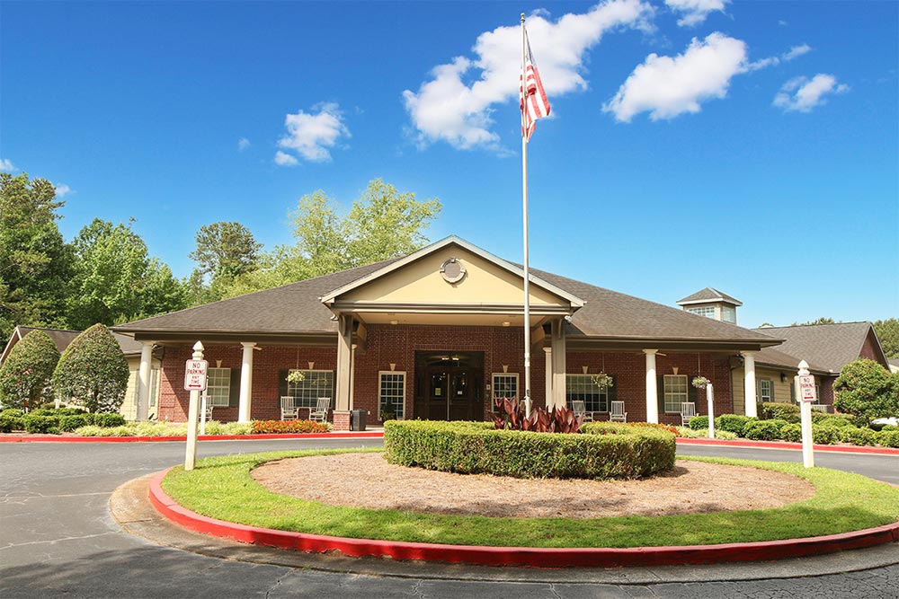 A brick building with a triangular roof entrance, an American flag on a flagpole, and a circular driveway bordered with red stands as a welcoming senior living residence in Illinois.