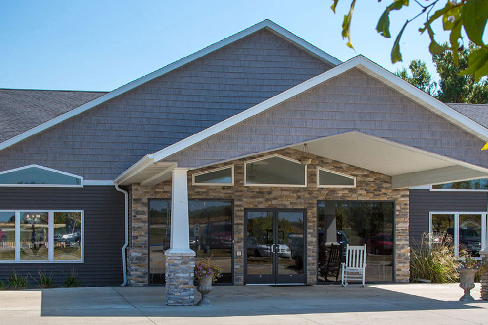 Single-story senior living facility in Illinois with a large covered entrance, double glass doors, stone accents, and a white rocking chair on the porch. The facade is clad in brown siding with a sloped roof.